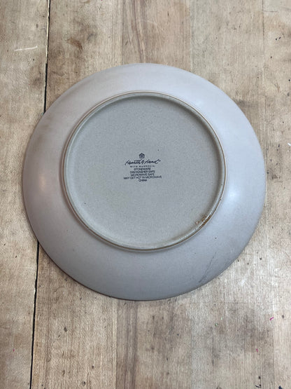 The underside of a round ceramic plate with a light gray glaze, featuring the printed text 'Royal Albert' and 'The Queen Mother Honored Edition 1974', placed on a rustic wooden surface.