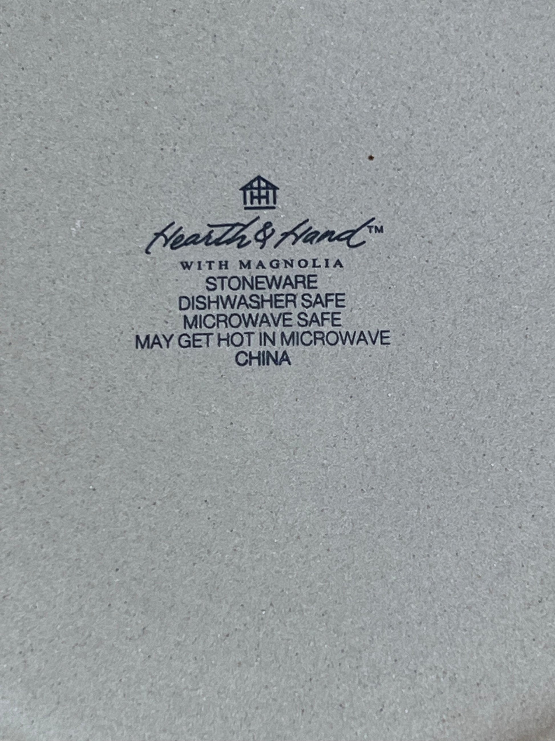 Close-up of a light gray stoneware surface with black printed text reading Heath & Hand with Magnolia, plus product details such as stoneware, dishwasher safe, microwave safe, and may get hot in microwave, China.