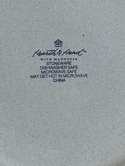 Close-up of a light gray stoneware surface with black printed text reading Heath & Hand with Magnolia, plus product details such as stoneware, dishwasher safe, microwave safe, and may get hot in microwave, China.