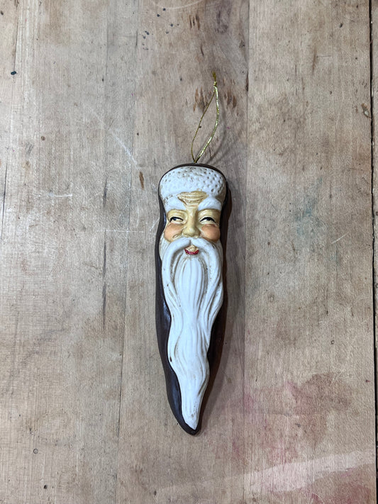 A hand-painted wooden ornament shaped like an elderly man's face with a long white beard and mustache, hanging on a rustic wooden plank background.