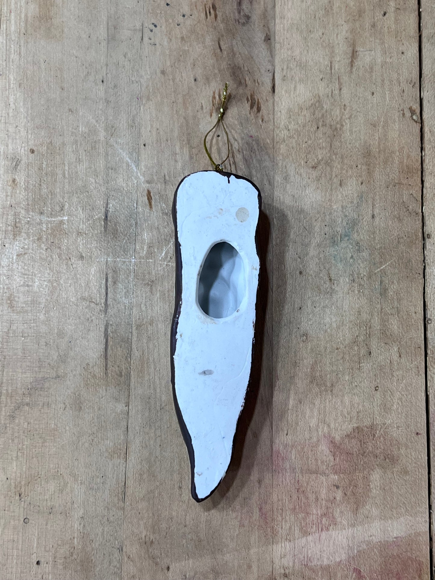 A rustic, handcrafted wooden ornament shaped like a chili pepper, featuring a white-painted center with a cutout, a dark brown wood edge, and a small gold metal hanging loop at the top, displayed on a light wood plank background.