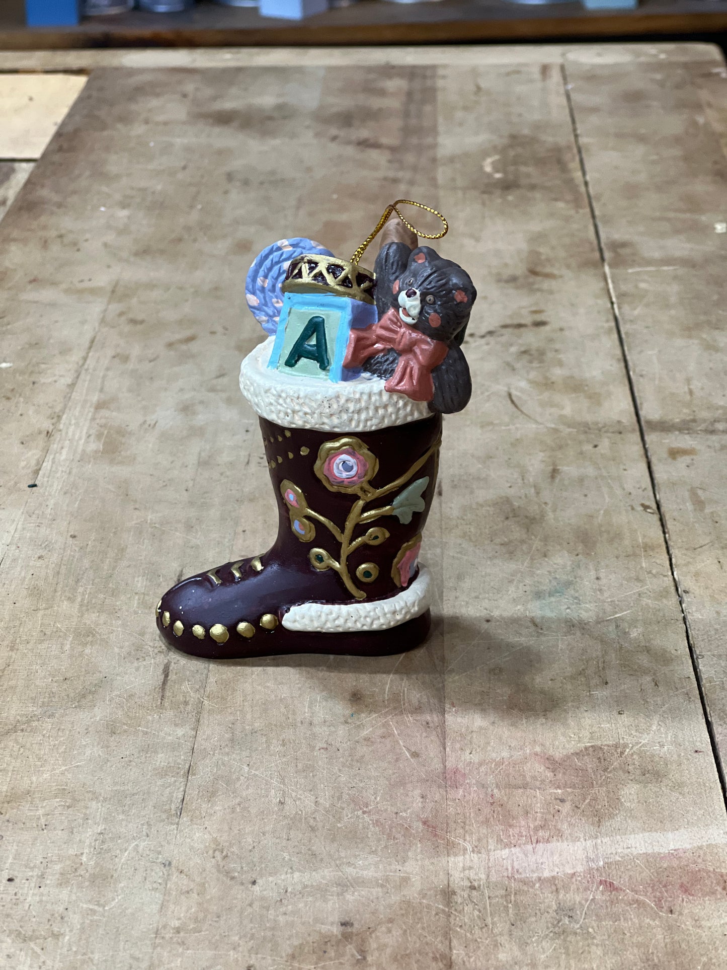 A decorative boot-shaped ornament with a dark brown body, white faux-fur trim, gold floral details, and a small hanging ornament on top featuring a gray cat, a blue letter A, and other holiday accents, displayed on a wooden surface.
