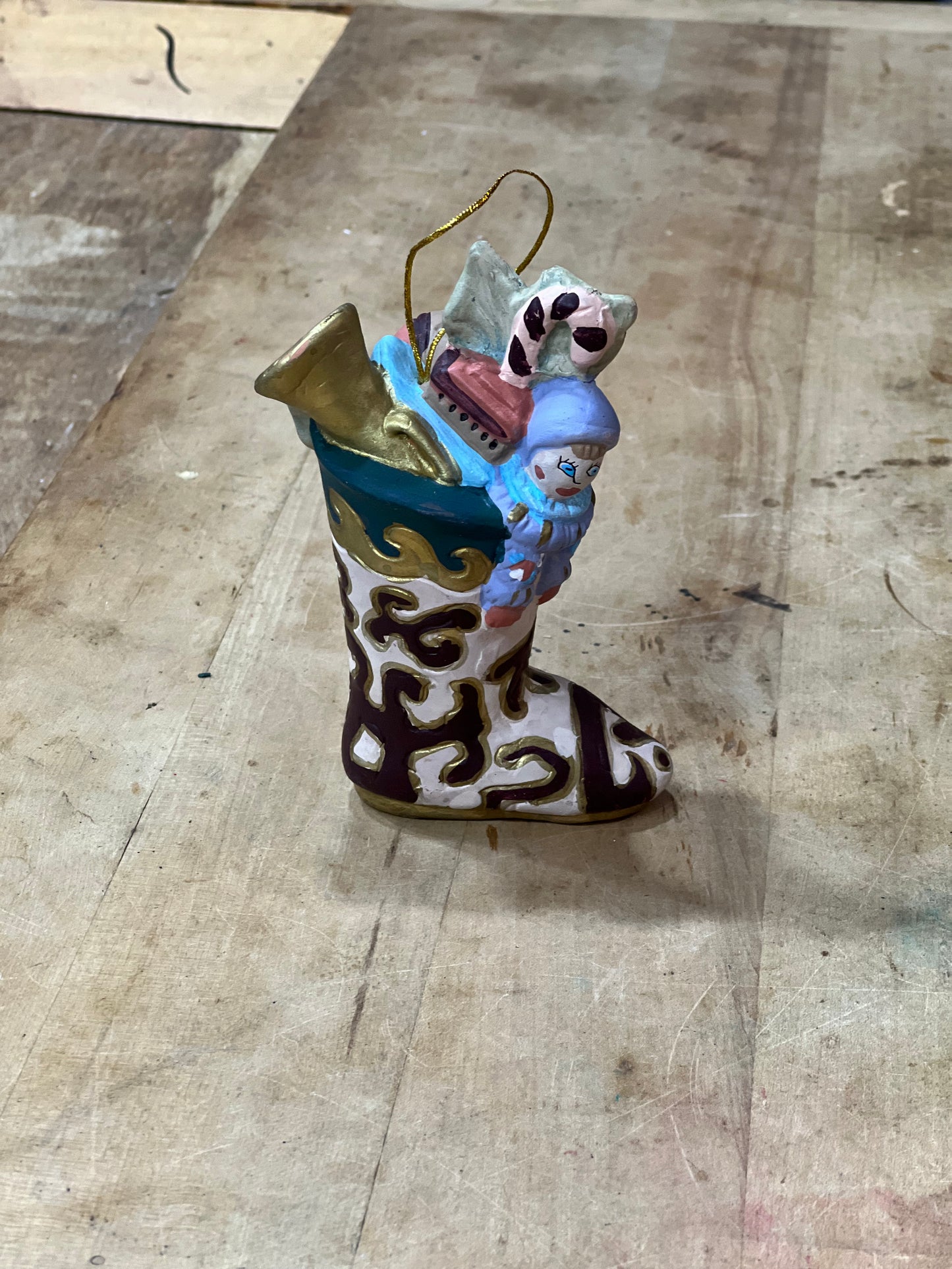 A small decorative ornament shaped like a white boot with dark brown abstract swirls, holding a blue snowman figure and gold holiday items, photographed on a worn wooden tabletop.