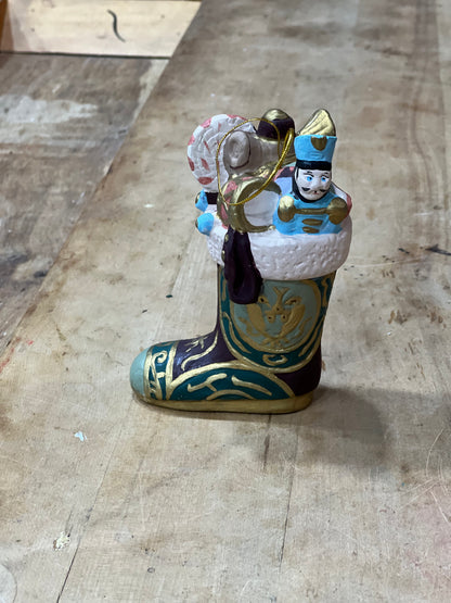 A decorative nutcracker figurine sitting inside a painted boot, placed on a worn wooden floor. The boot is decorated with teal, gold, and dark purple patterns, and the nutcracker wears a blue uniform with gold accents.