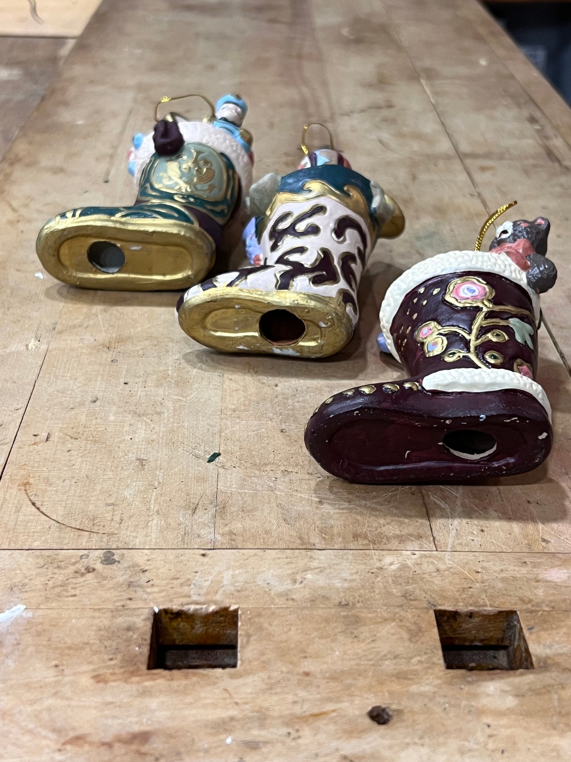 Three hand-painted ceramic Chinese New Year ornaments shaped like boots, displayed on a wooden workbench.