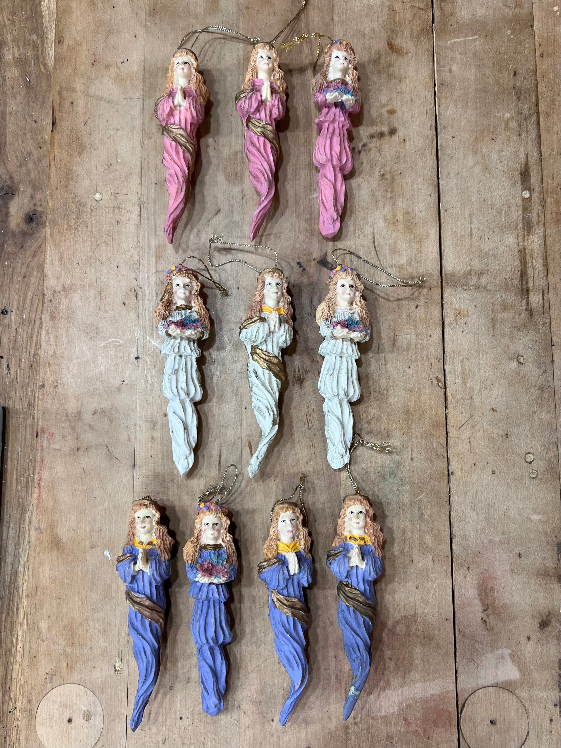 Nine small hanging figurines or ornaments arranged on a rustic wooden surface. Each figure has a painted face and draped body in pink, white, or blue tones, with some wearing gold rope details and small multicolored floral accents near the waist.