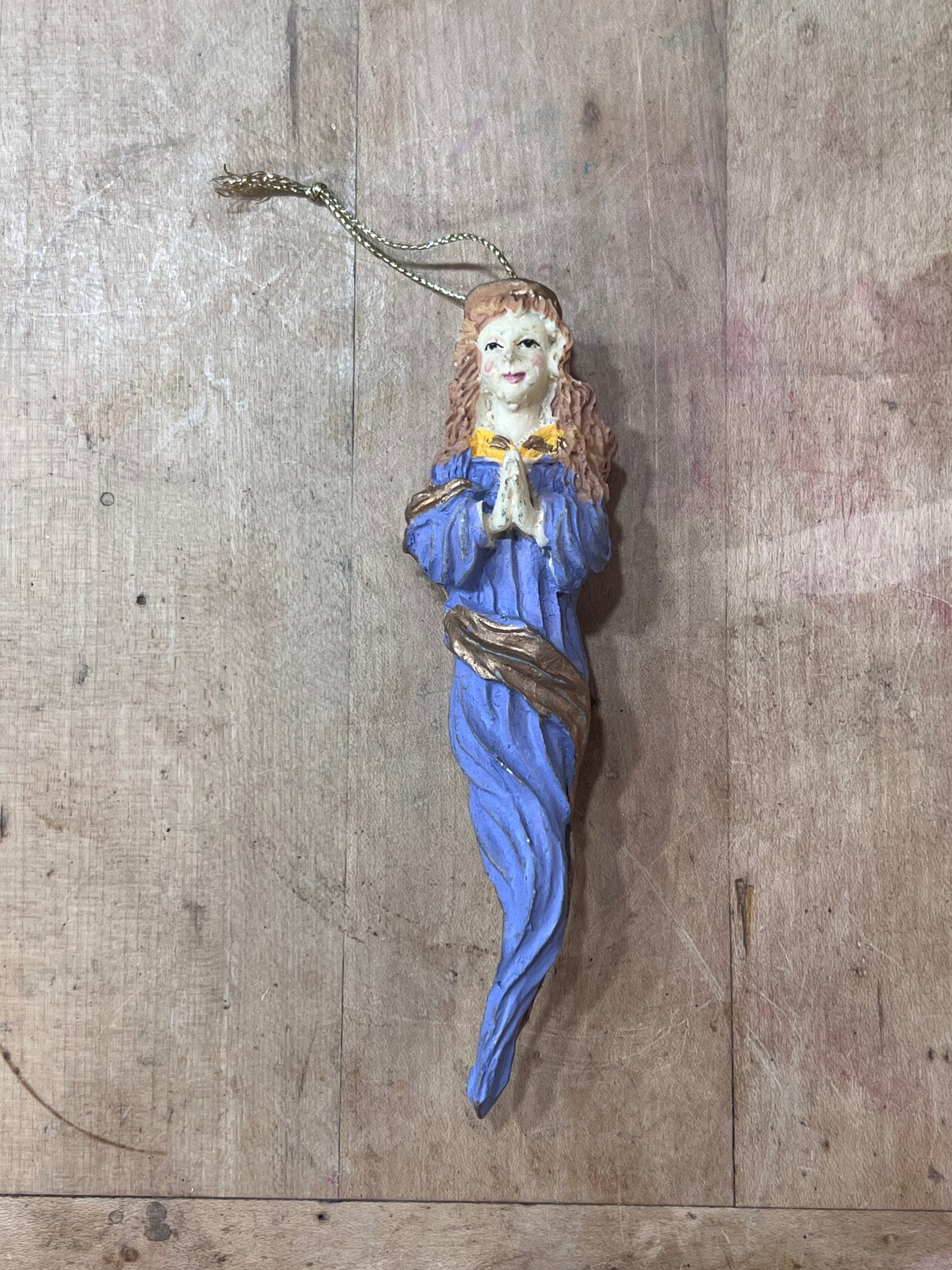 A small hand-painted Christmas ornament shaped like a praying woman in a blue robe with a gold collar, hanging from a gold chain against a rustic wooden background.