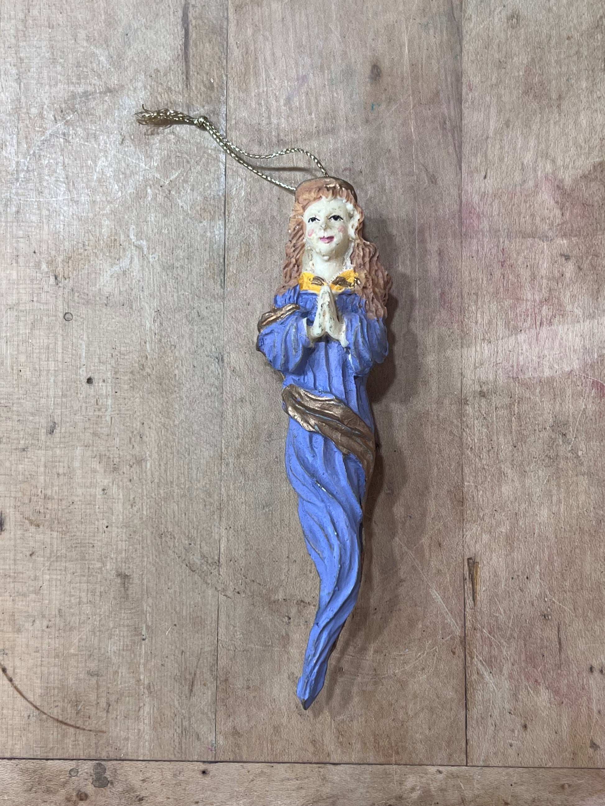 A small hand-painted Christmas ornament shaped like a praying woman in a blue robe with a gold collar, hanging from a gold chain against a rustic wooden background.