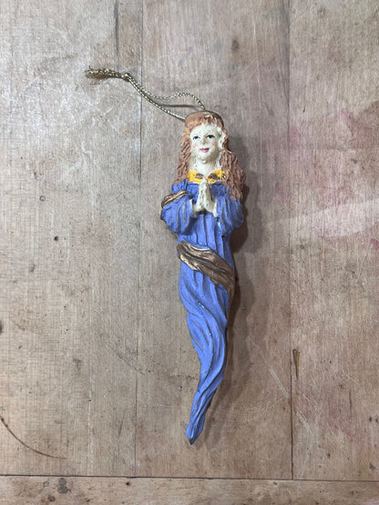 A small hand-painted Christmas ornament shaped like a praying woman in a blue robe with a gold collar, hanging from a gold chain against a rustic wooden background.