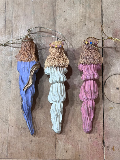 Three Christmas tree ornaments shaped like pinecones with textured brown tops and hanging loops, shown in blue, white, and pink on a rustic wooden background.