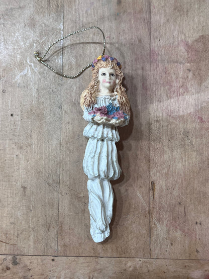 A vintage-style resin Christmas ornament shaped like a young girl with long wavy light brown hair, wearing a white dress and holding a small bouquet of blue and pink flowers. The ornament hangs from a silver-colored metal chain against a rustic wooden background.