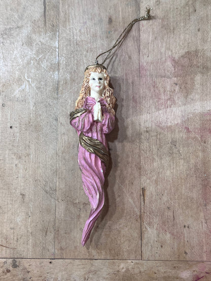 A vintage-style hanging ornament depicting a praying angel or woman in a pink robe with gold accents, shown against a rustic wooden plank background.