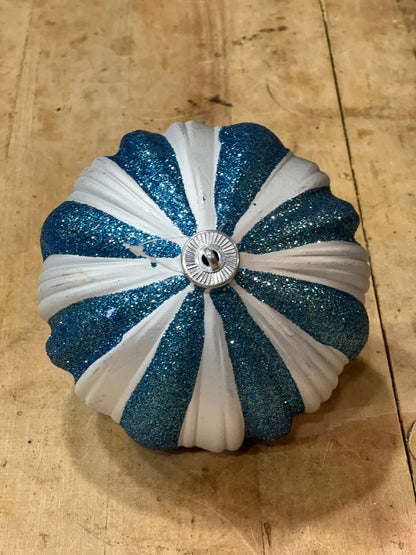A round, ribbed ornament or decorative piece with alternating glittery blue and white stripes, featuring a silver metallic button in the center, resting on a light wood surface.