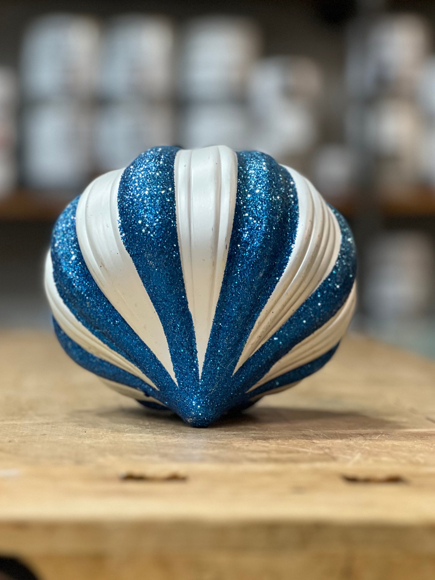 A seashell-shaped decorative ornament with alternating glittery blue and matte white ridges, resting on a wooden surface with a blurred background.