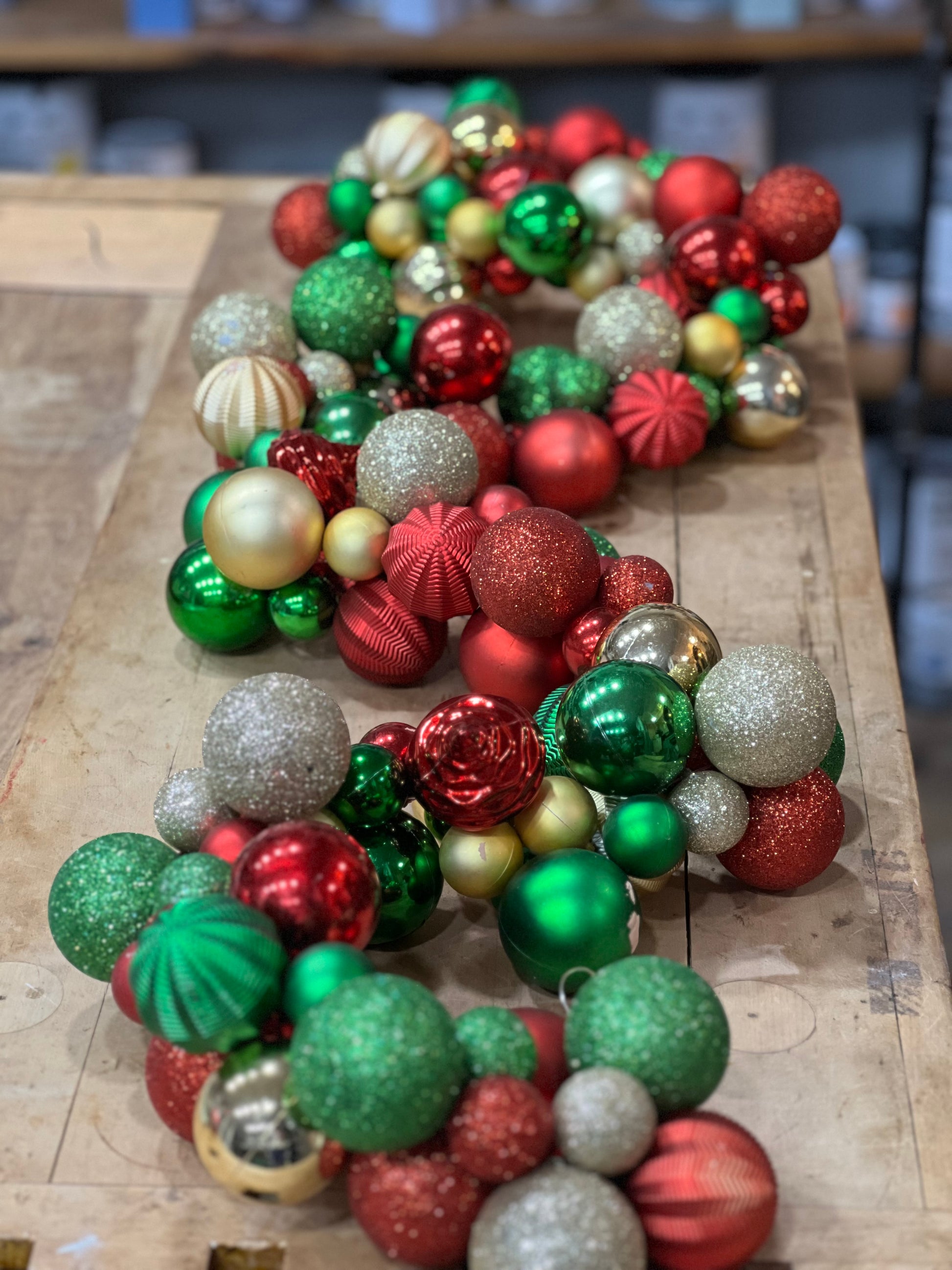 A collection of assorted Christmas ornaments in red, green, silver, and gold, including matte, glitter, and ribbed styles, arranged on a rustic wooden surface.