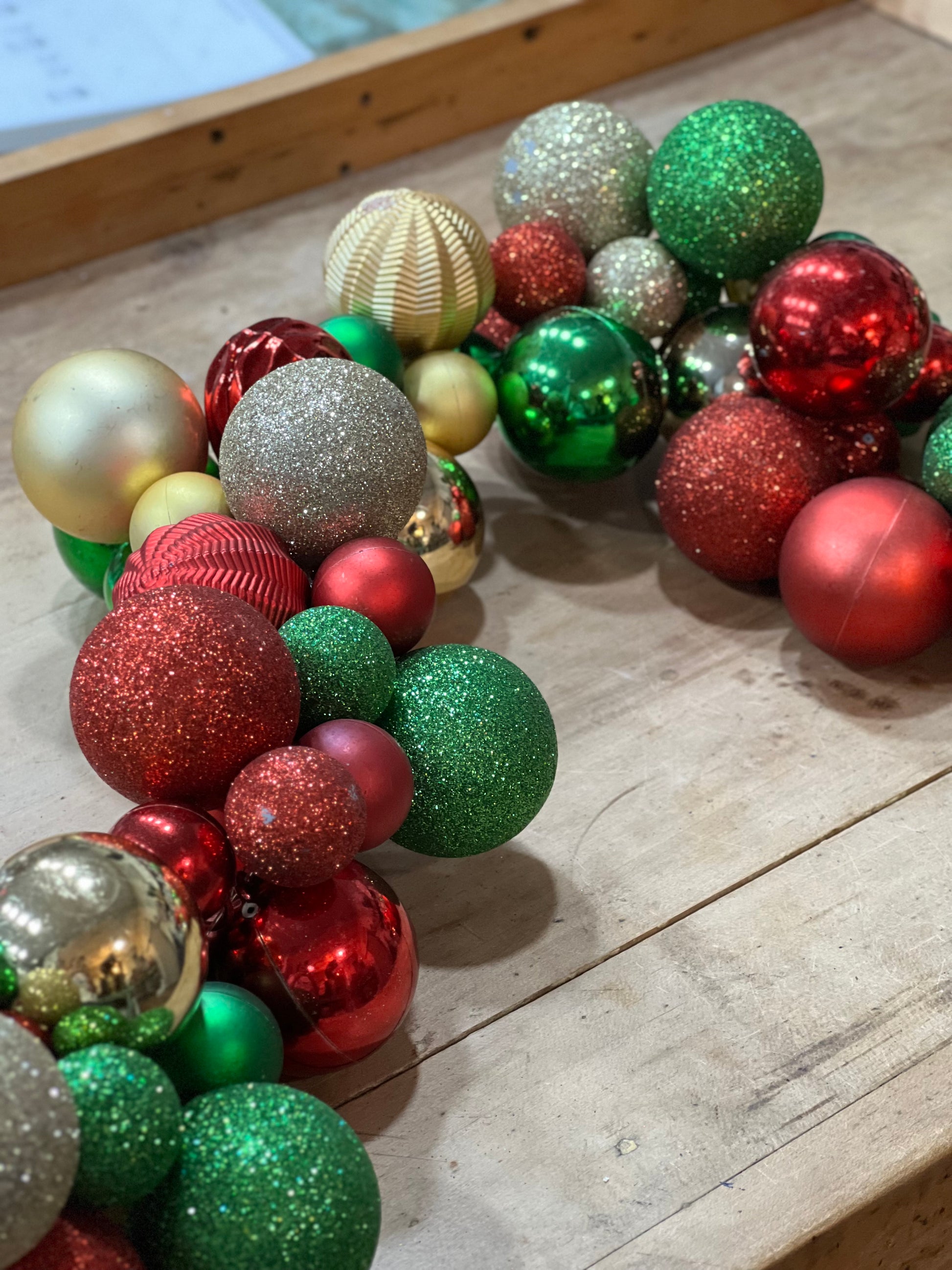 A collection of assorted Christmas ornaments, including red, green, silver, and gold baubles with glitter and ribbed textures, arranged on a light-colored wooden surface.