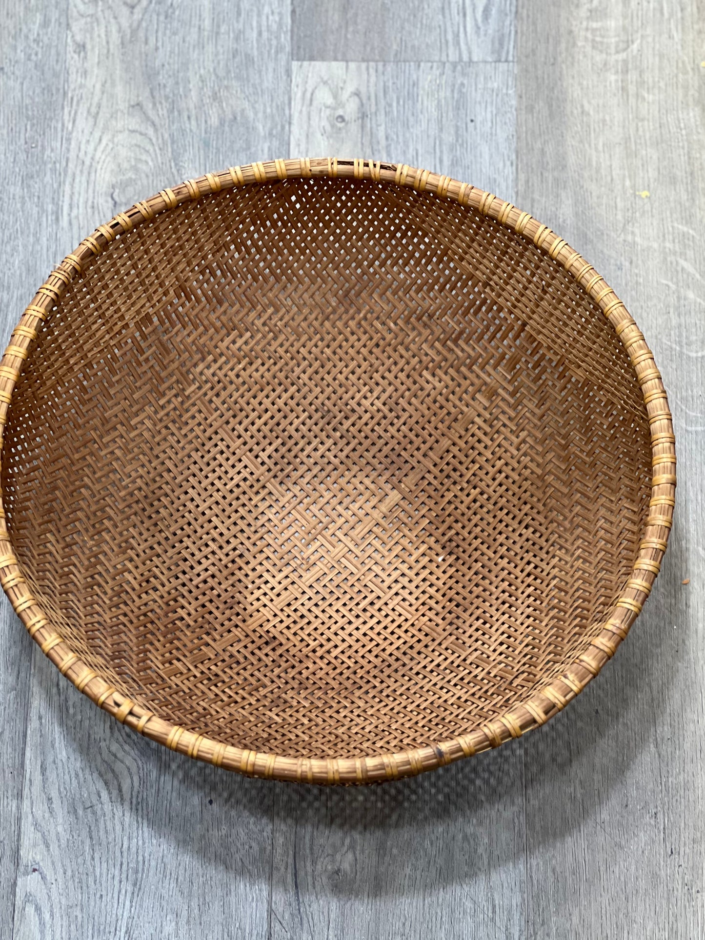 A round woven rattan basket with a natural brown color, photographed from a top-down perspective on a gray wood-look floor.