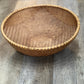 A round woven natural fiber basket with a hand-woven rim, shown on a gray wood plank floor.