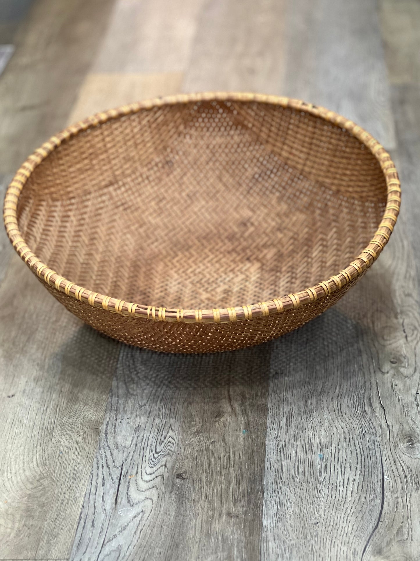 A round woven natural fiber basket with a hand-woven rim, shown on a gray wood plank floor.