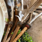 A close-up of several decorative feather quills with ribbed wooden handles and metallic accents, arranged with small green foliage on a rustic wooden surface.