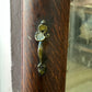 An ornate vintage-style metal door pull handle with a distressed bronze and gold finish, mounted on a dark brown wooden door frame.
