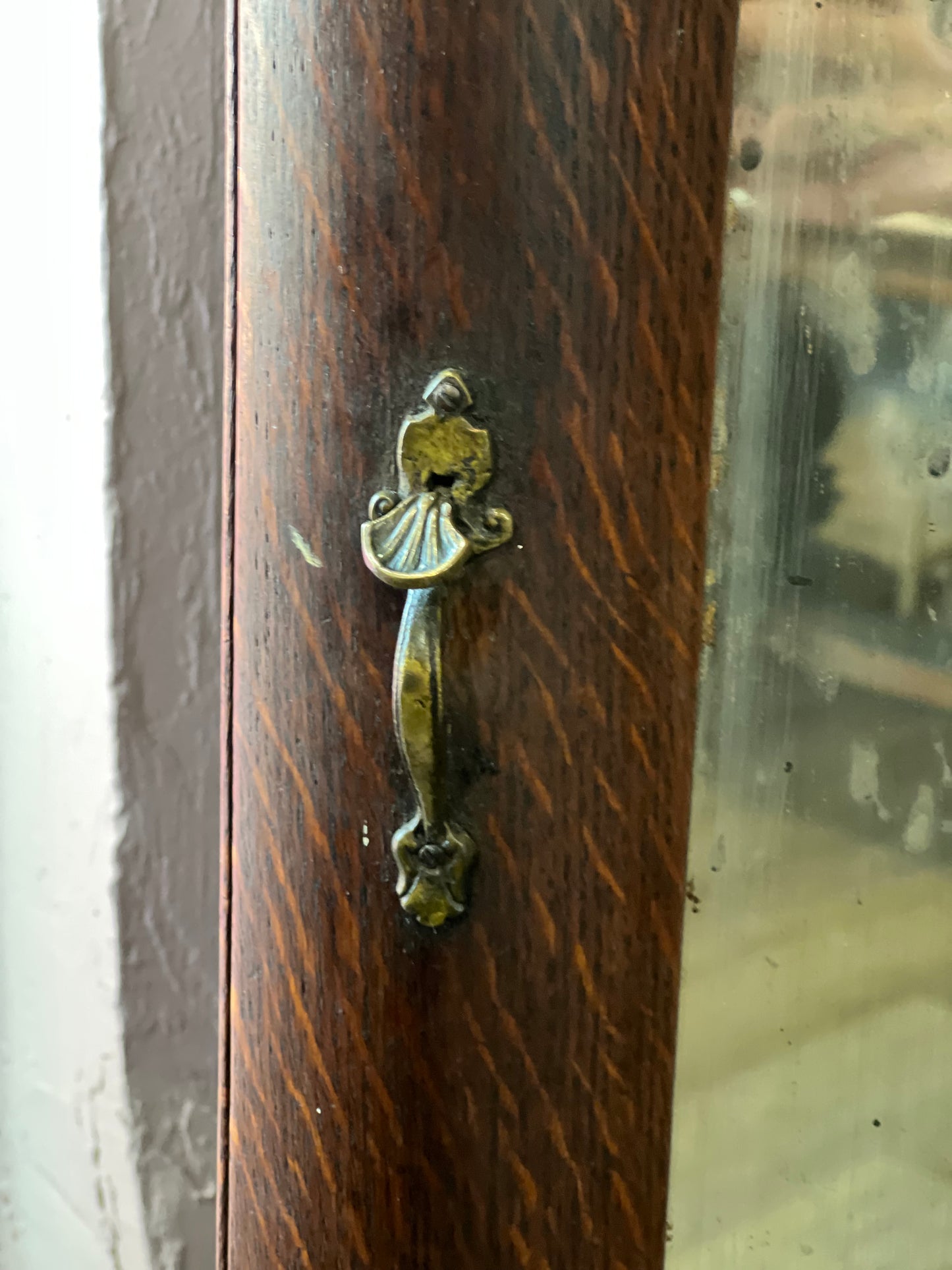 An ornate vintage-style metal door pull handle with a distressed bronze and gold finish, mounted on a dark brown wooden door frame.
