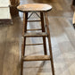 A rustic, vintage wooden step stool with a round seat and three steps, standing on a light brown wooden floor in what appears to be a shop or home interior.