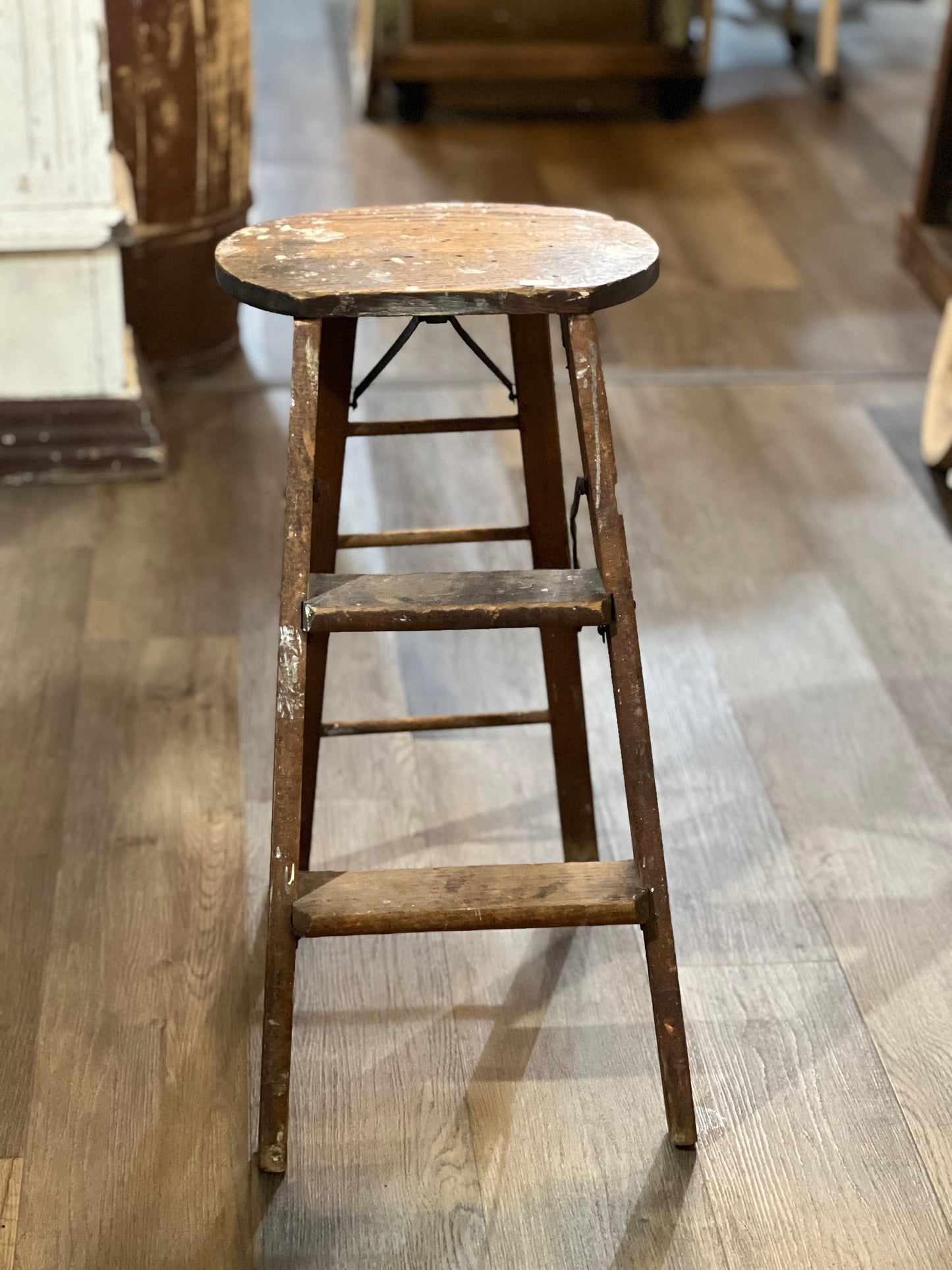 A rustic, vintage wooden step stool with a round seat and three steps, standing on a light brown wooden floor in what appears to be a shop or home interior.