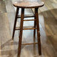 A rustic wooden bar stool with a round seat and ladder-style legs, shown on a light wood floor.