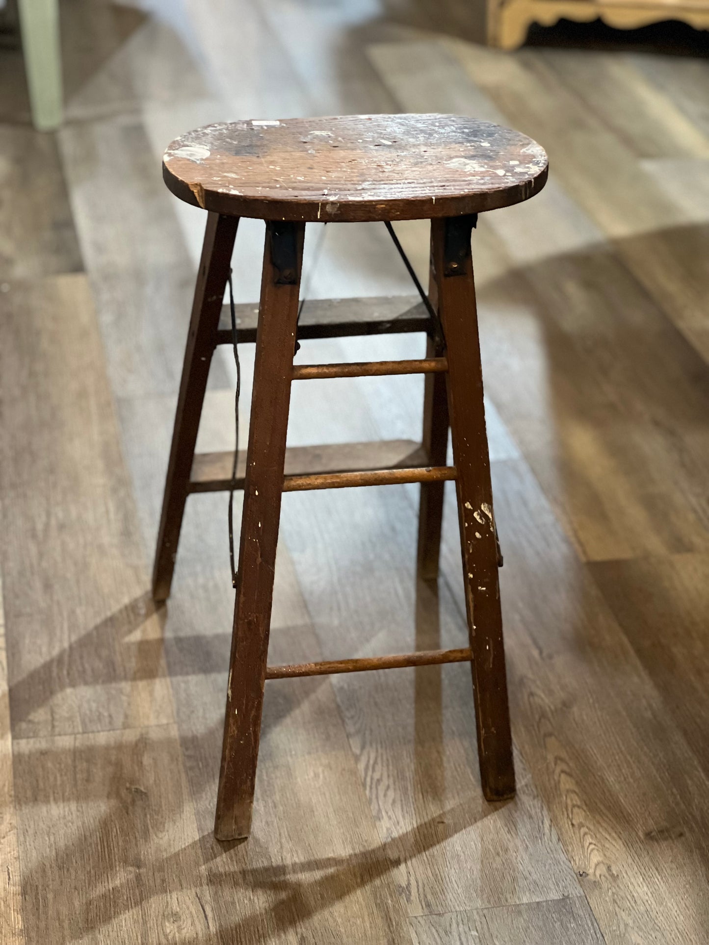 A rustic wooden bar stool with a round seat and ladder-style legs, shown on a light wood floor.