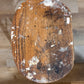 A top-down view of a round wooden stool or stool seat with a heavily distressed finish, showing peeling white paint, scratches, and dark stains on a light brown wood floor.