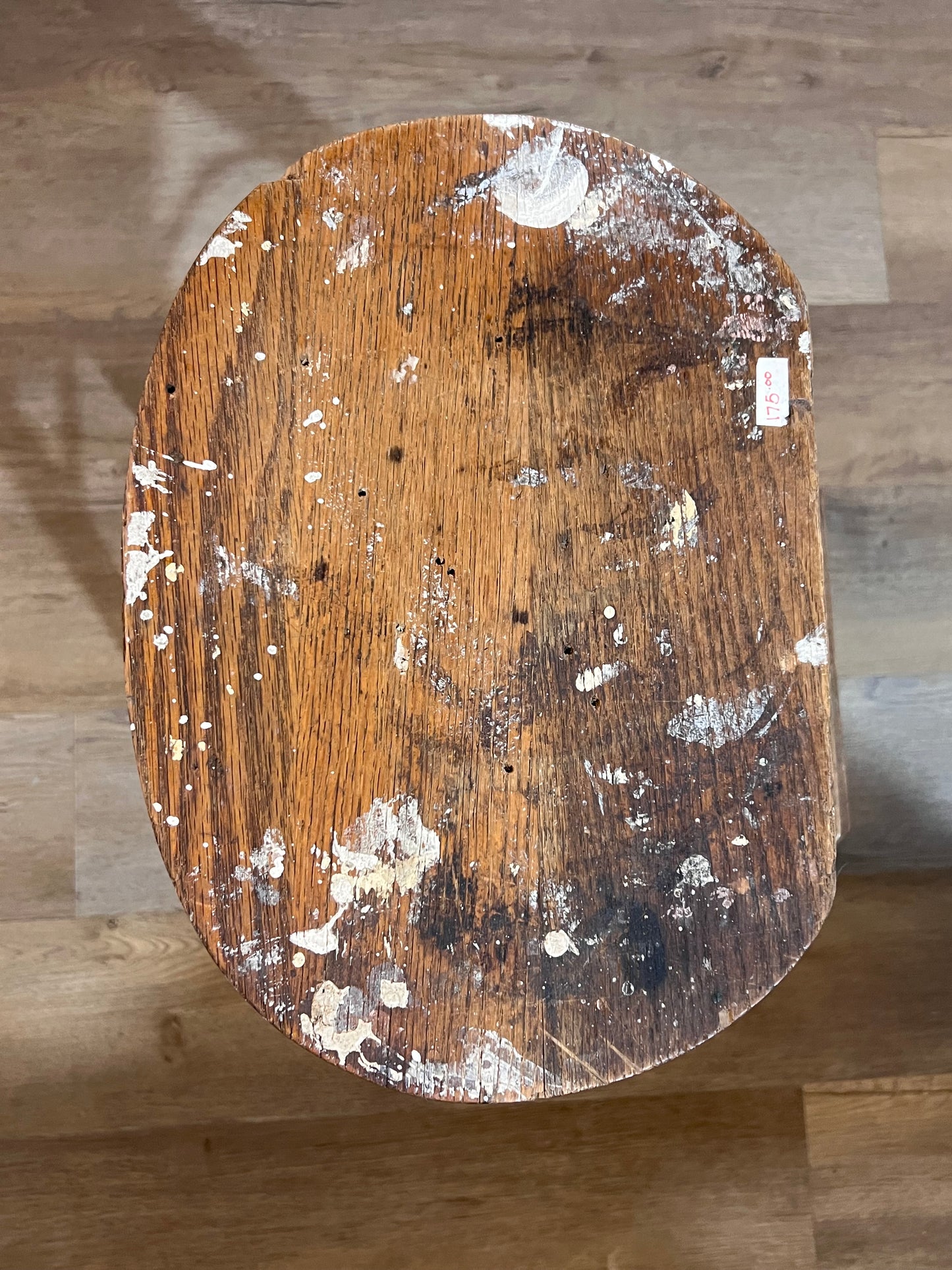 A top-down view of a round wooden stool or stool seat with a heavily distressed finish, showing peeling white paint, scratches, and dark stains on a light brown wood floor.