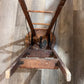 An antique wooden stool with a round seat and four legs, photographed upside down on a light brown wood plank floor.