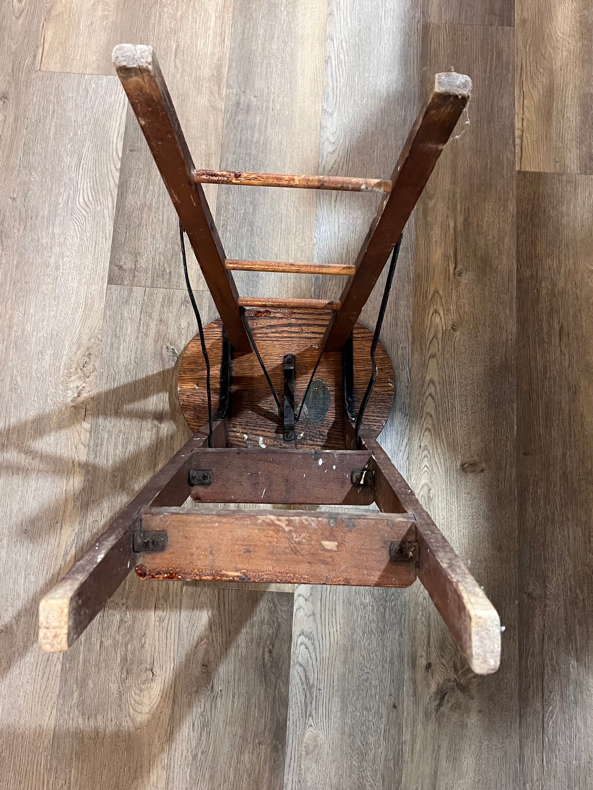 An antique wooden stool with a round seat and four legs, photographed upside down on a light brown wood plank floor.
