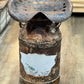 A rustic, vintage-style metal pedestal stool made from a heavily rusted milk can with a perforated metal seat on top, set on a light wood floor.
