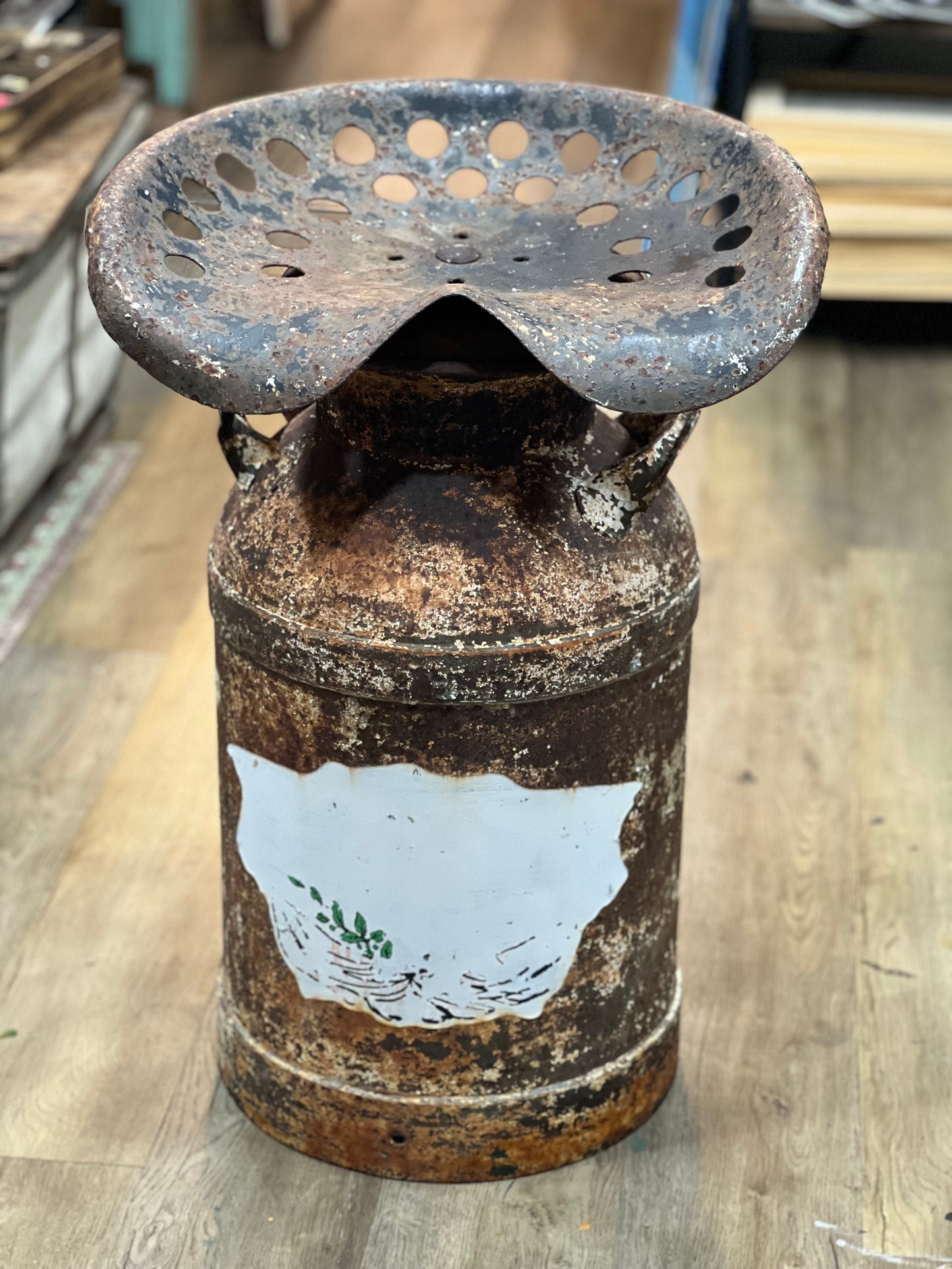 A rustic, vintage-style metal pedestal stool made from a heavily rusted milk can with a perforated metal seat on top, set on a light wood floor.