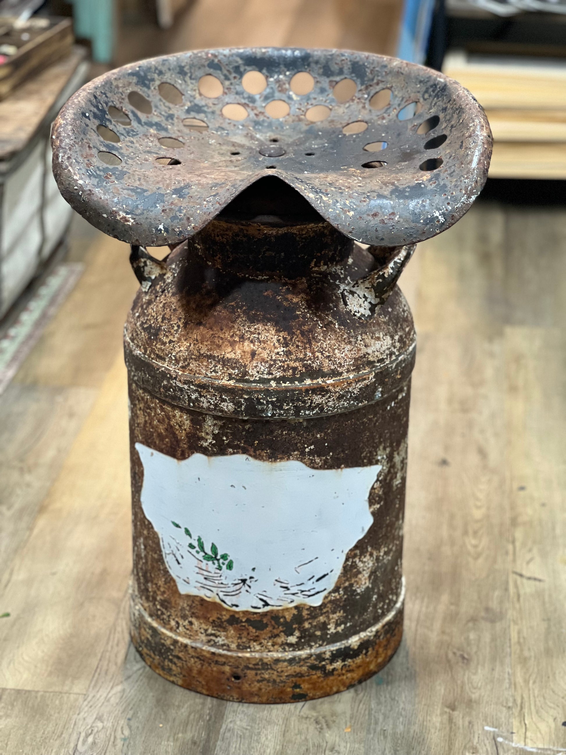A rustic, vintage-style metal pedestal stool made from a heavily rusted milk can with a perforated metal seat on top, set on a light wood floor.