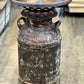A distressed, vintage-style rusted metal milk can or decorative container with a perforated lid, standing on a light wood floor.