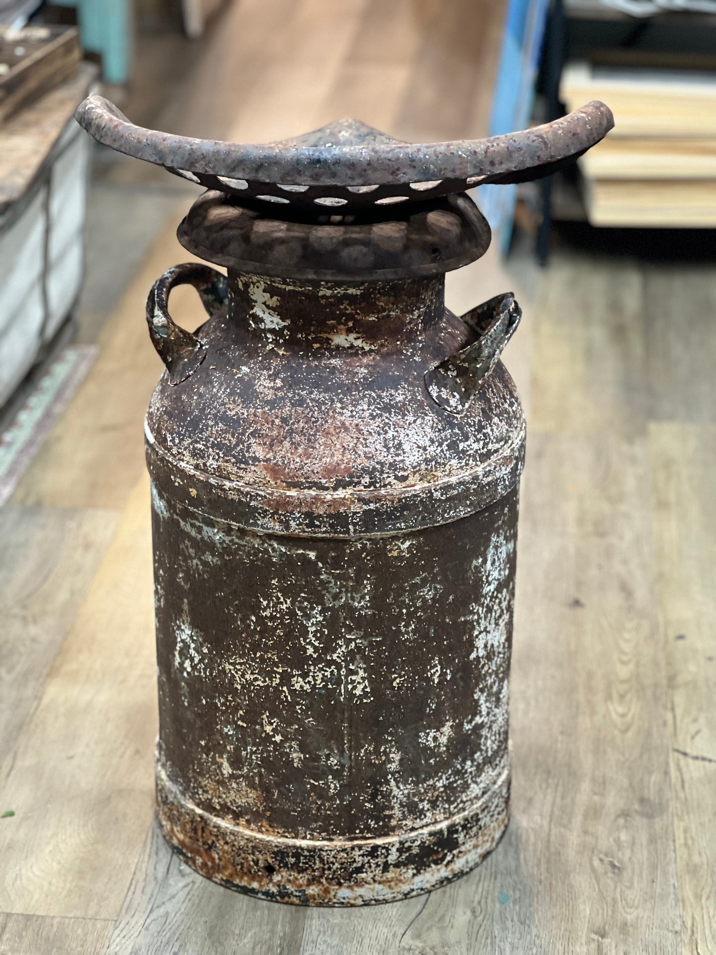 A distressed, vintage-style rusted metal milk can or decorative container with a perforated lid, standing on a light wood floor.