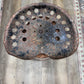 A rustic, heavily weathered metal stool seat with multiple circular cutouts, showing significant rust and patina, photographed from above on a wood-look floor.