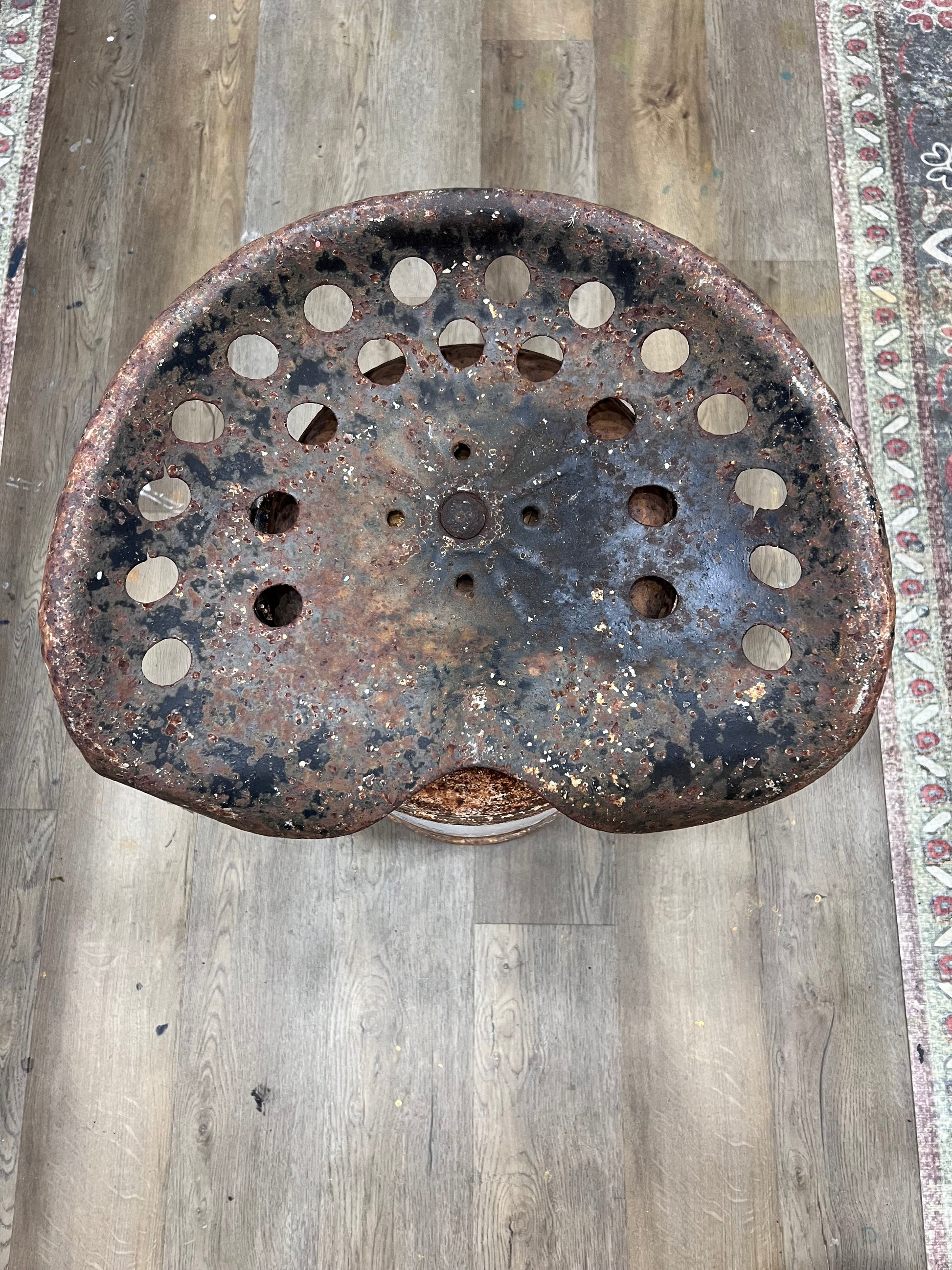 A rustic, heavily weathered metal stool seat with multiple circular cutouts, showing significant rust and patina, photographed from above on a wood-look floor.