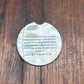A round coasters with a cutout for a drink, featuring a printed Bible verse from Matthew 17:10 and a botanical design, displayed on a dark wood background.