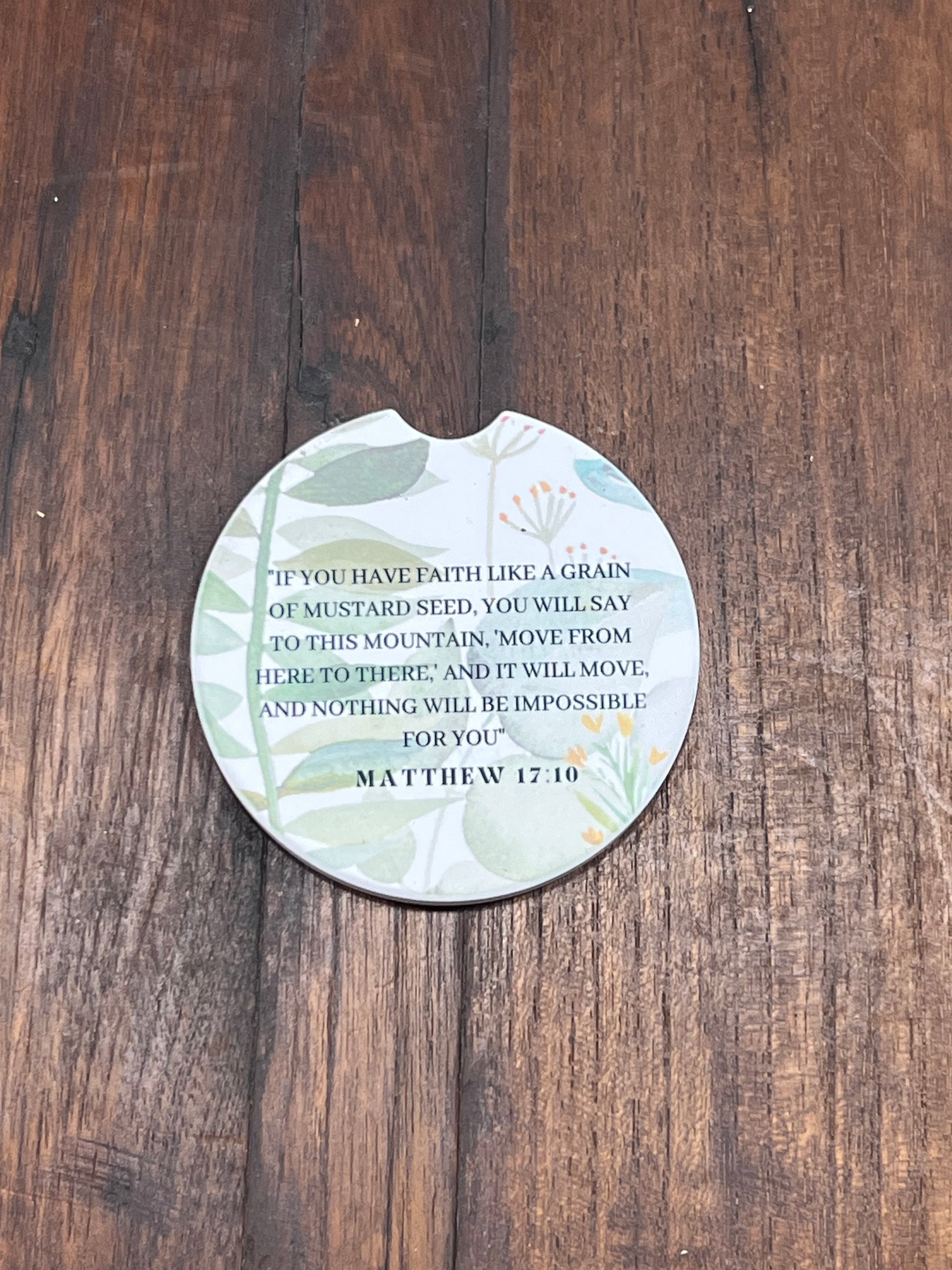 A round coasters with a cutout for a drink, featuring a printed Bible verse from Matthew 17:10 and a botanical design, displayed on a dark wood background.