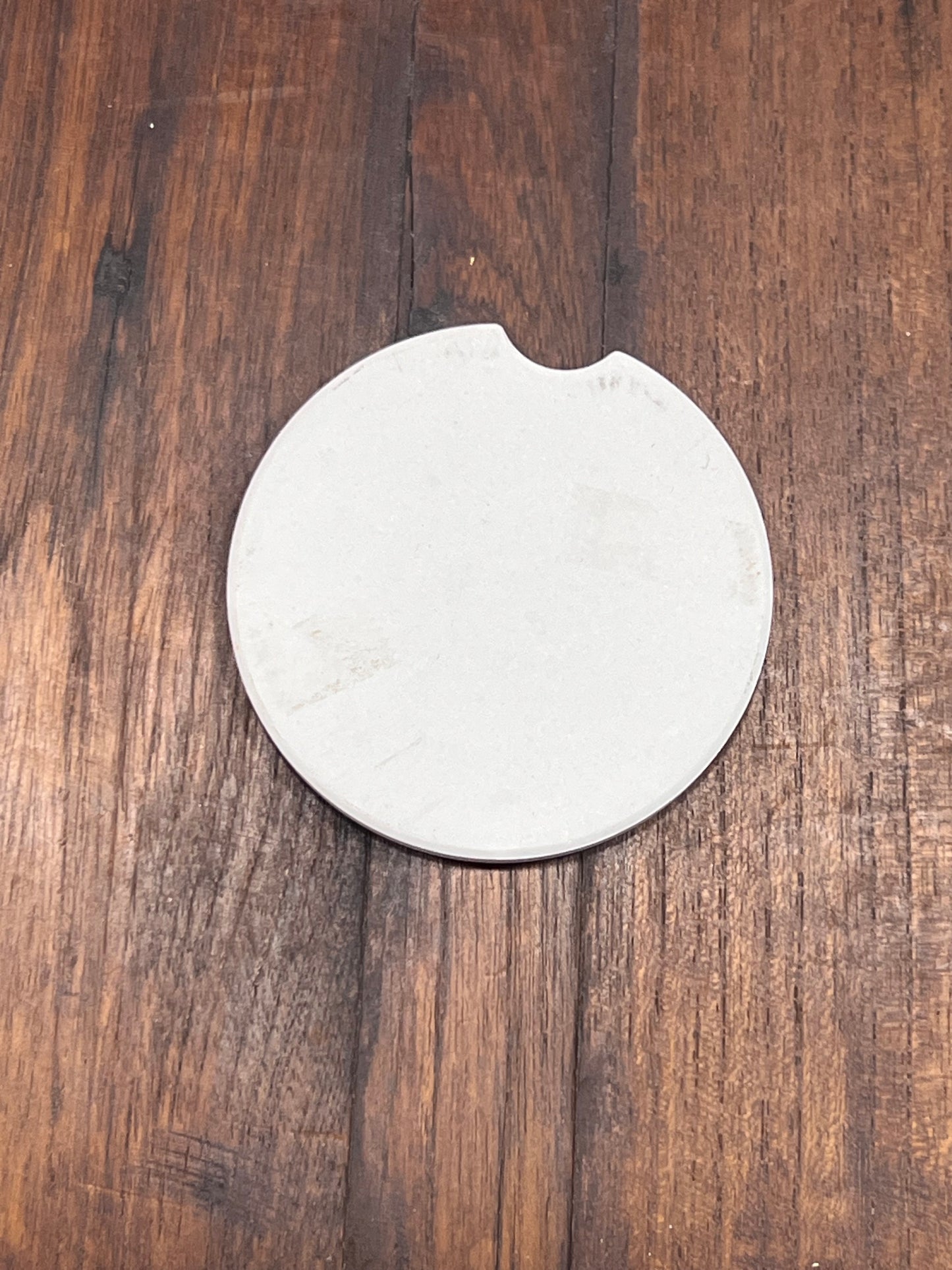 A round off-white ceramic coaster with a small notch on the top edge, placed on a dark brown wooden surface.