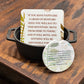 A square printed card with a Bible verse sits in a small black metal pot, with a round matching pin or sticker placed in front on a rustic wooden surface.