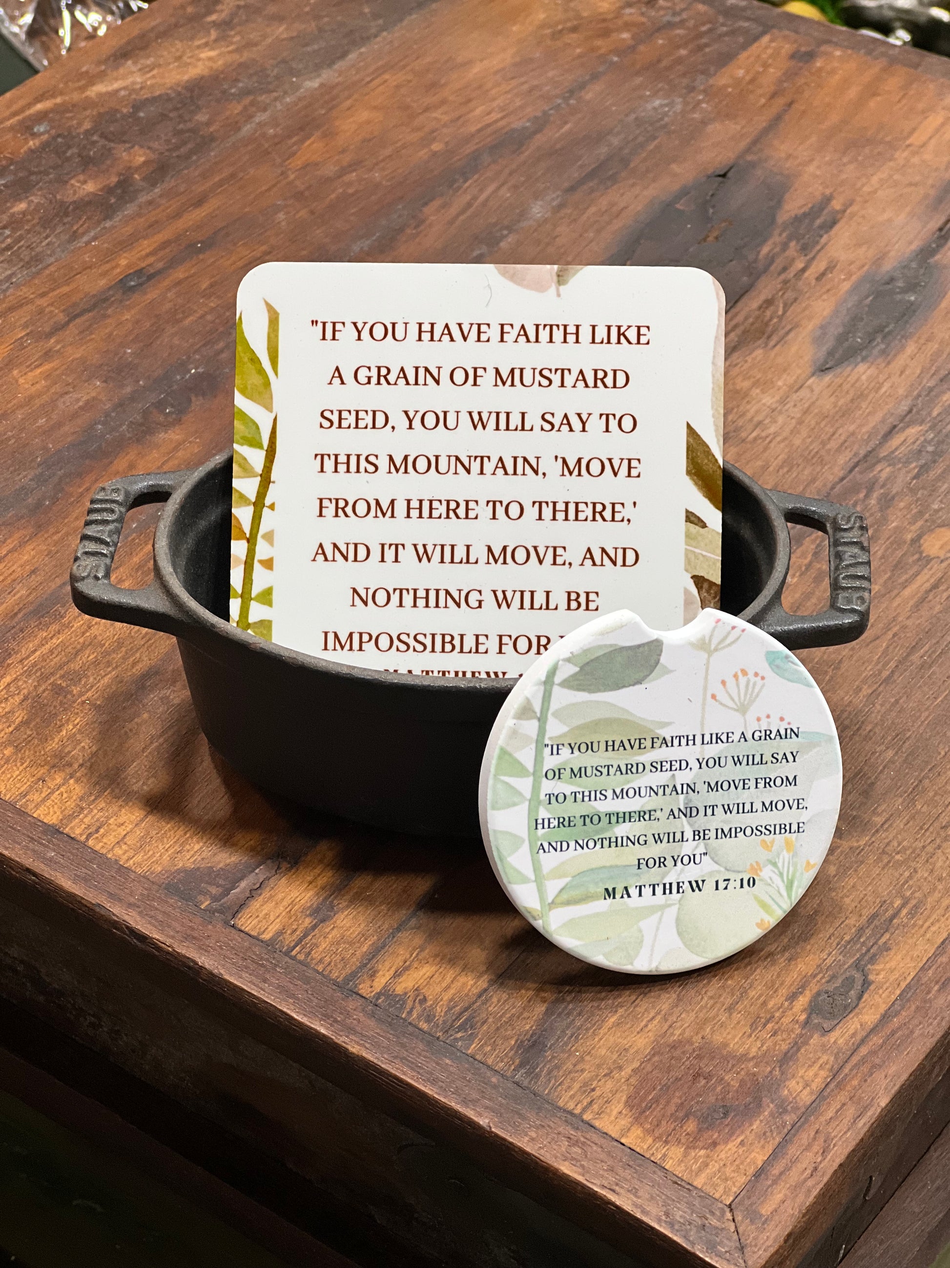A square printed card with a Bible verse sits in a small black metal pot, with a round matching pin or sticker placed in front on a rustic wooden surface.