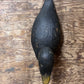 A hand-painted wooden duck deco, likely a mallard duck, featuring a dark black-brown body with a natural wood grain texture and a yellow-tipped beak, displayed on a rustic wooden surface.