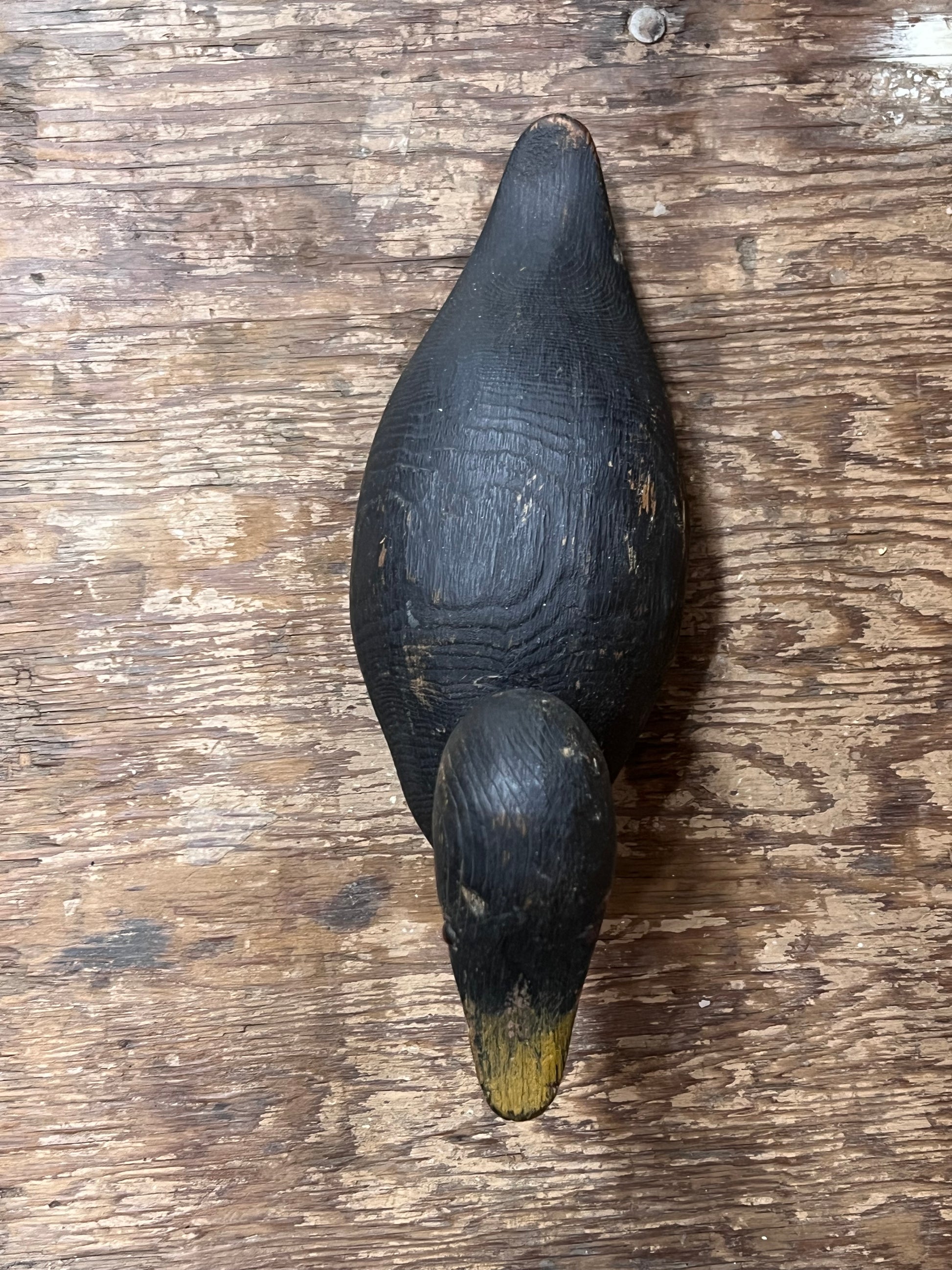 A hand-painted wooden duck deco, likely a mallard duck, featuring a dark black-brown body with a natural wood grain texture and a yellow-tipped beak, displayed on a rustic wooden surface.