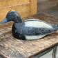 A hand-painted wooden duck deco shaped like a shoveler's duck, featuring a black head and back with a white belly, resting on a rustic wooden table.