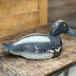 A hand-painted wooden duck deco, likely a tufted duck, resting on a rustic wooden table. The deco features a black head and back, a white belly, and a small yellow eye with a gray beak.